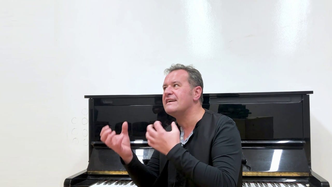 Robert Koziel speaking in front of a piano in I Know My Way